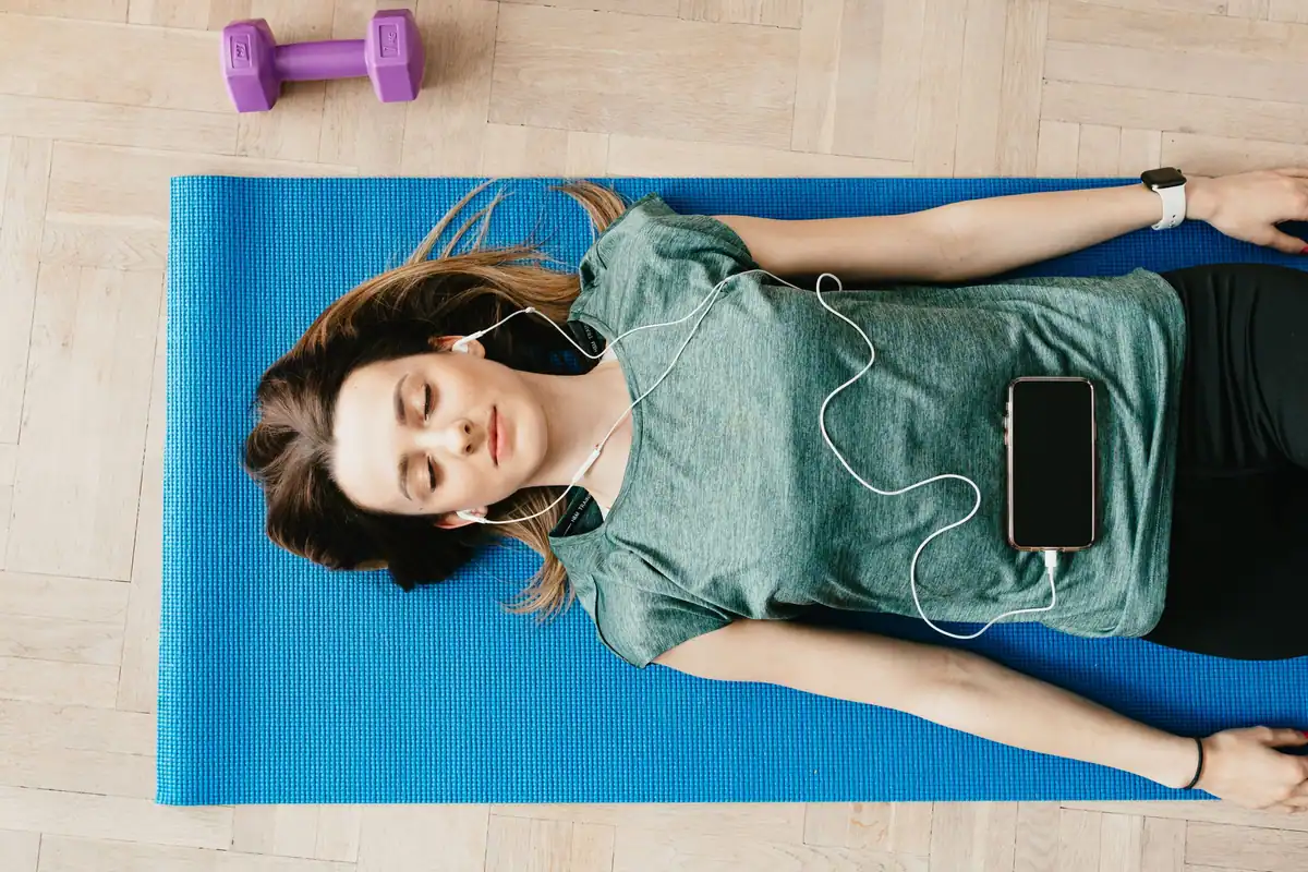 Tânără întinsă pe saltea de yoga care ascultă în căști o sesiune de pe telefon