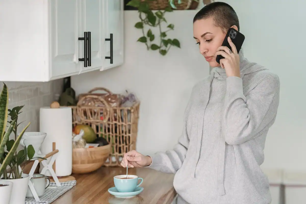 Femeie calmă în haine de casă care vorbește la telefon și amestecă în cafea în bucătărie