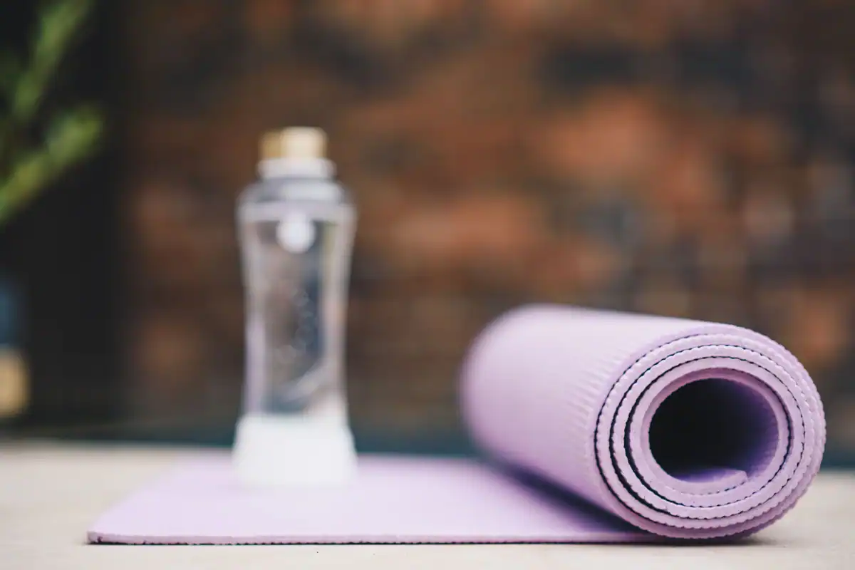 Saltea de yoga violet rulată lângă o sticlă de apă transparentă