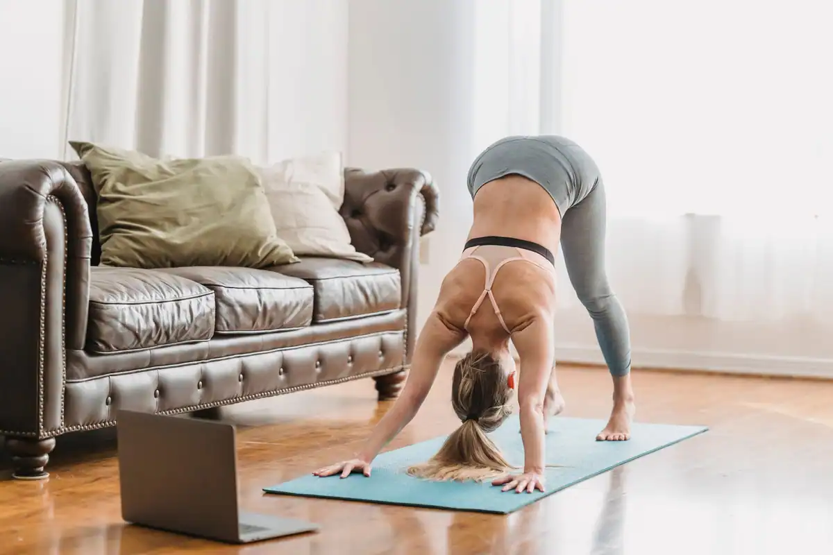 Femeie în colanți care se întinde pe saltea de yoga lângă laptop, în sufrageria luminoasă