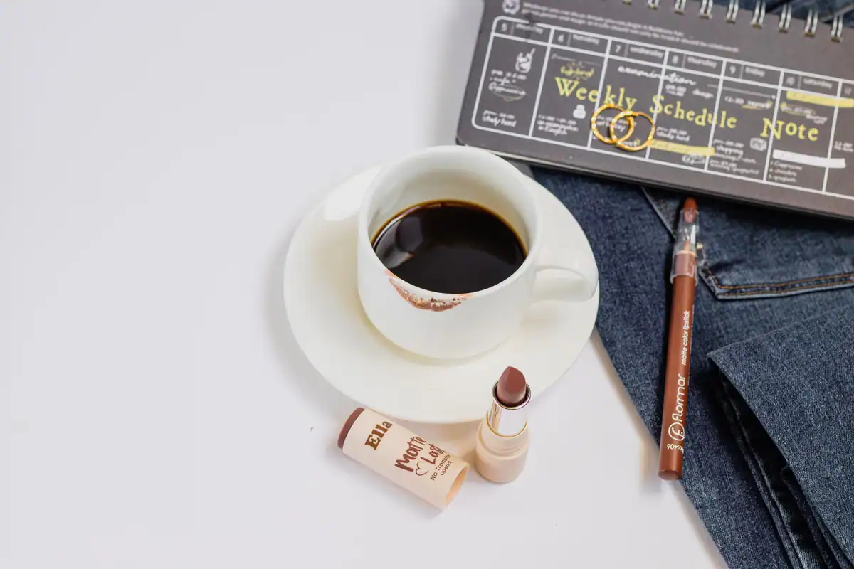 Agendă de planificare, cafea, ruj și blugi așezate pe masă, văzute de sus